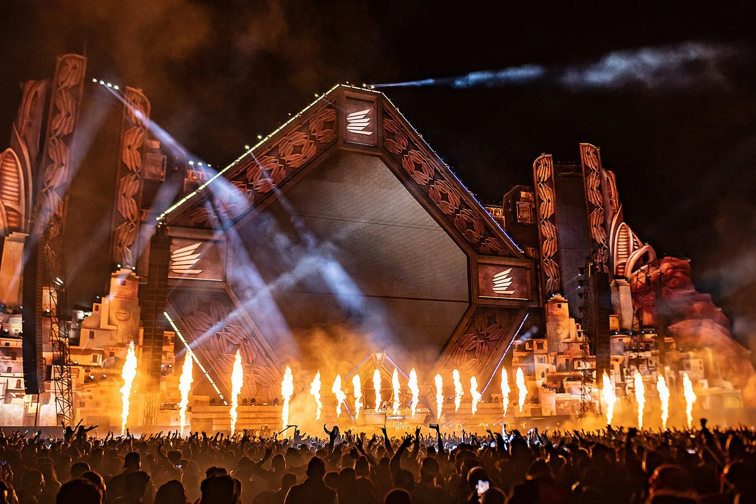 A large outdoor concert stage at night with fireworks and pyrotechnics, a crowd of people with raised hands, and colorful lights illuminating the scene.