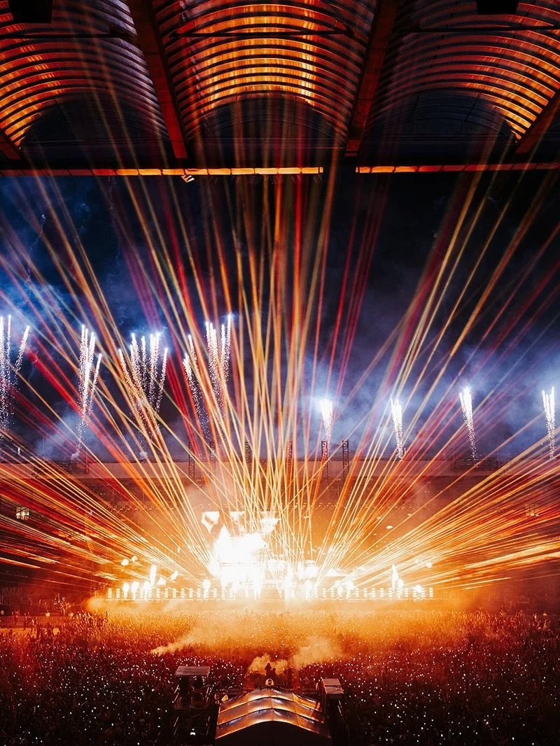 A large concert with fireworks, laser lights, and bright flames lighting up a crowd at night.