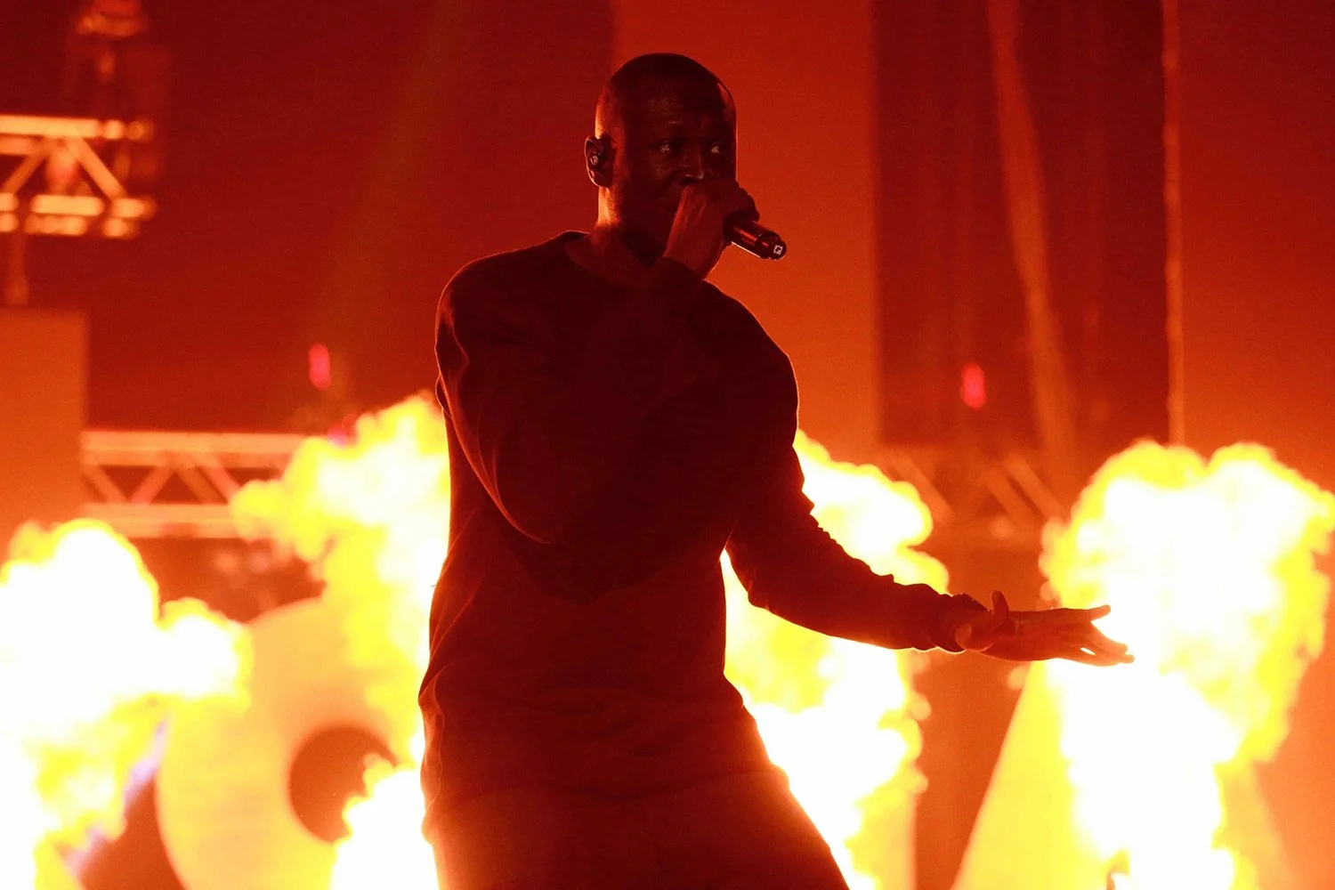 A male performer holding a microphone on stage with flames behind him during a concert.