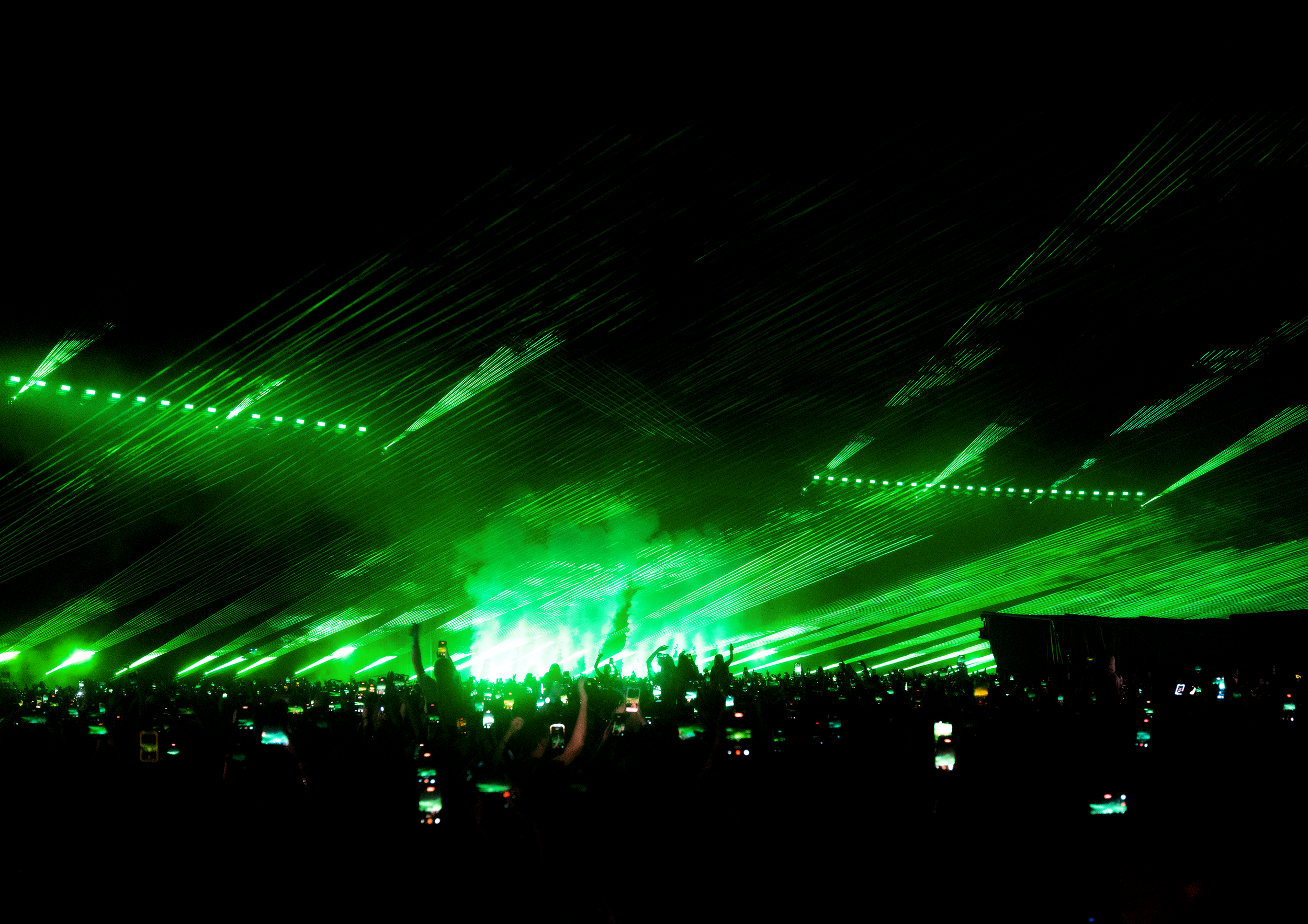 A concert scene with bright green laser lights and many people in the audience taking photos.