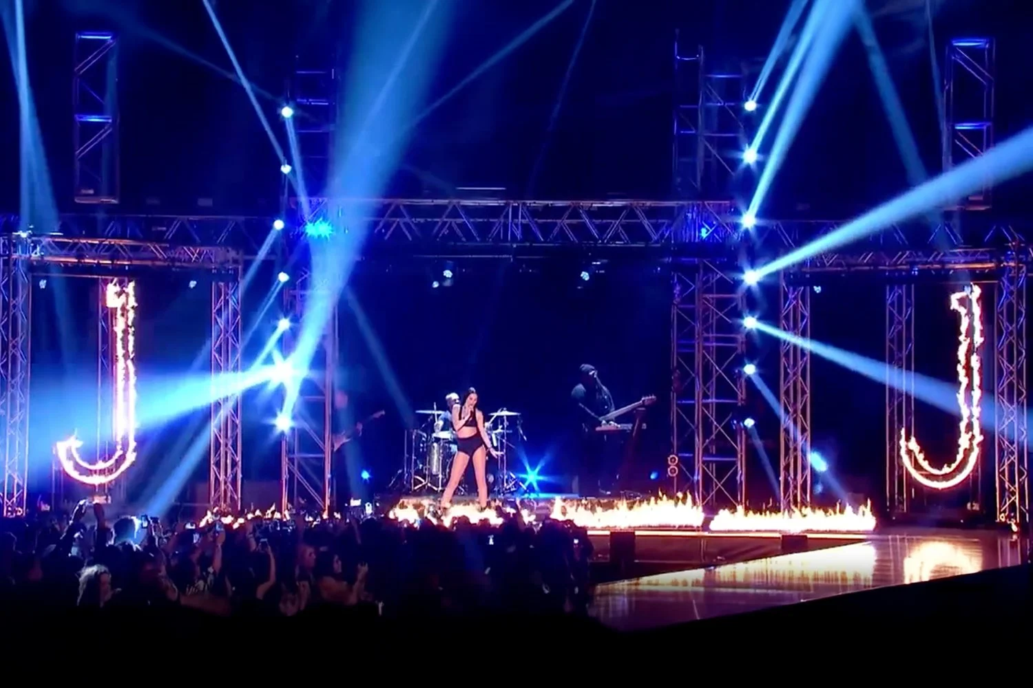 Stage with a singer in black outfit performing, surrounded by bright blue and white stage lights, with a band playing background and fire effects on the floor, audience watching in front.