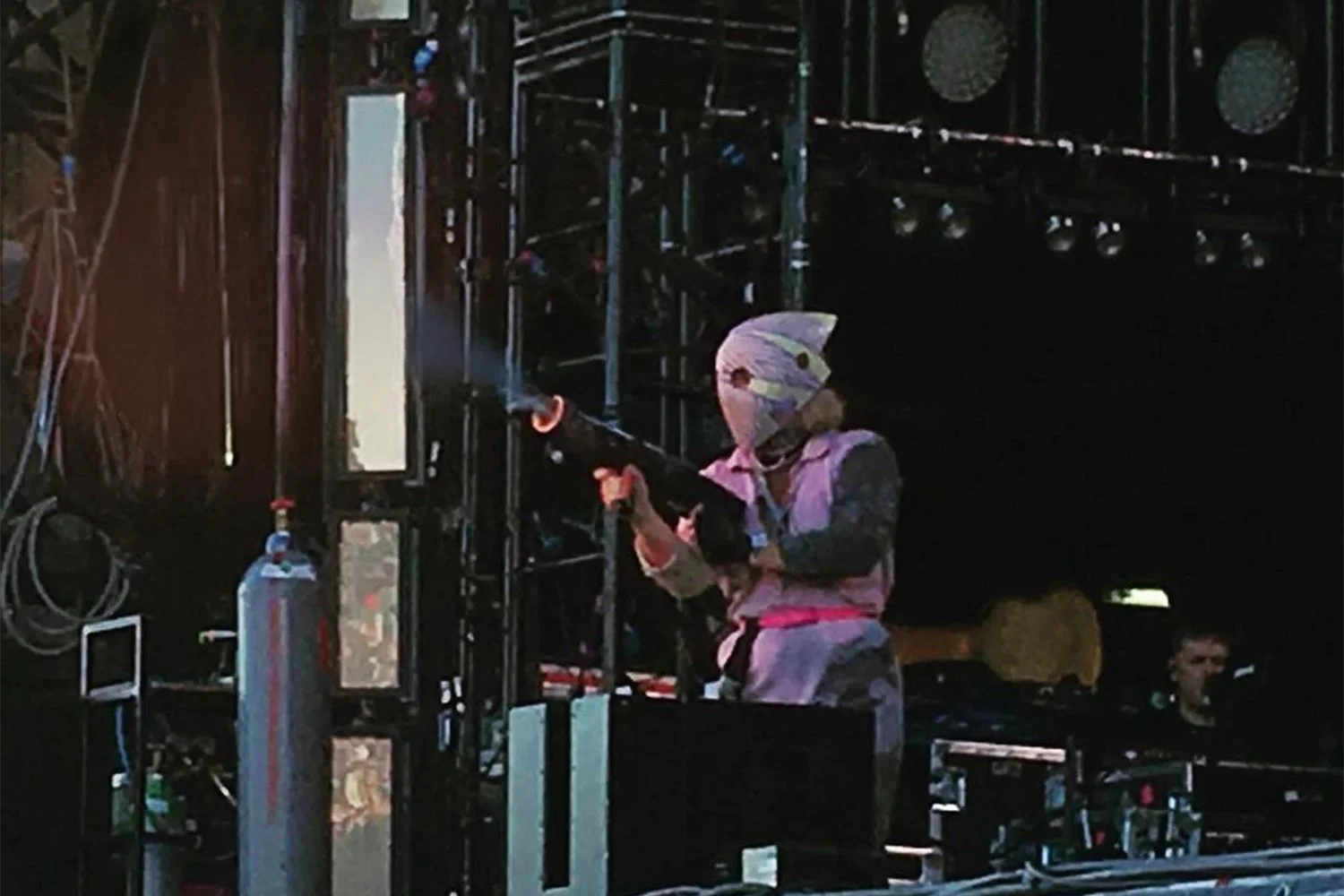 A person in a helmet and winter clothing aiming a gun with a scope in an indoor or stage setting with scaffolding and equipment.