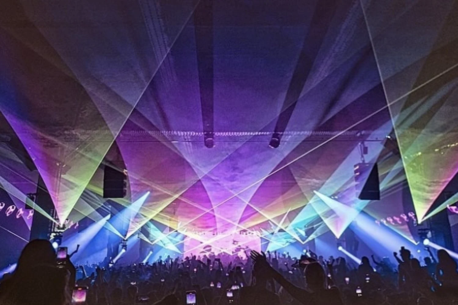 A concert or music event with colorful laser lights, a large crowd, and some attendees taking pictures with their phones.