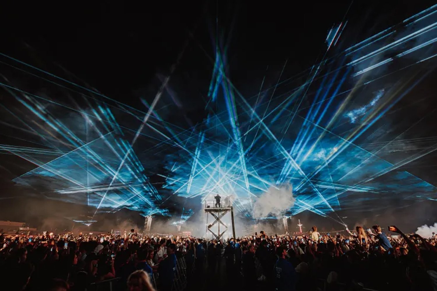 A large concert or festival with a crowd of people watching a laser light show at night. The show features blue laser beams and smoke effects above the stage.