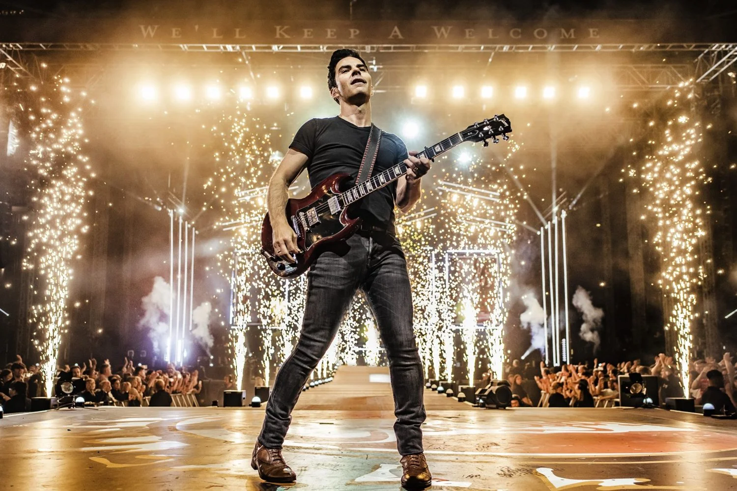 A male musician on stage playing an electric guitar during a concert with a backdrop of fireworks and bright lights, with an audience watching.