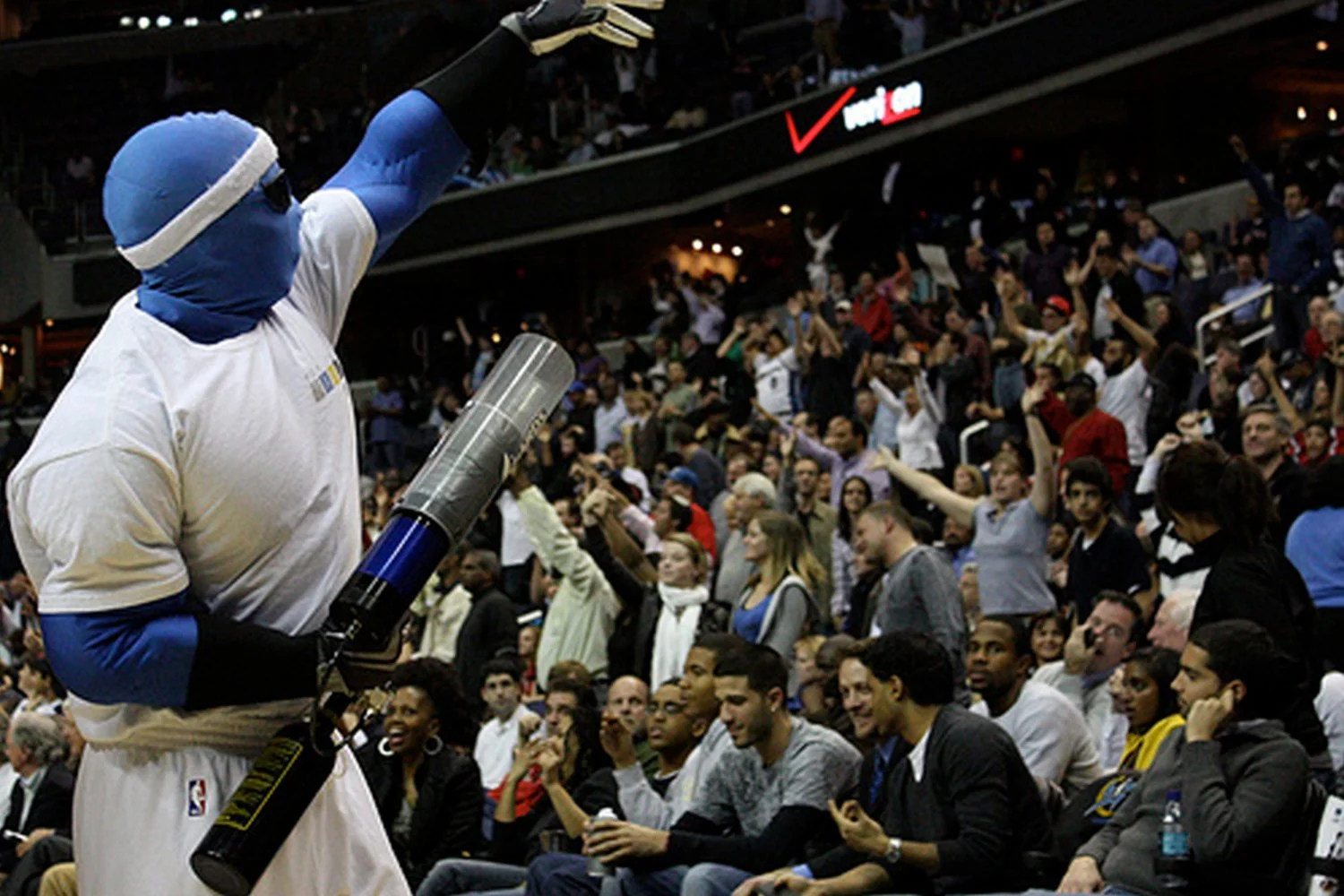 A person dressed in a blue costume and white basketball jersey holding a large novelty rocket prop, raising their arm to a crowd of excited spectators at a sports arena.