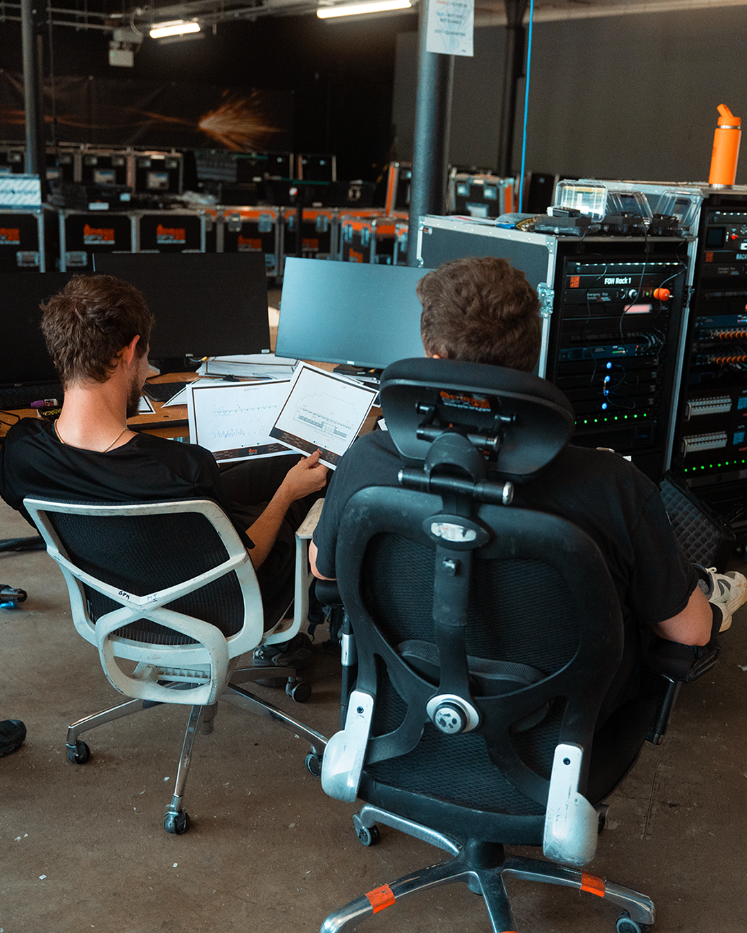 Two people sitting at a desk with multiple monitors and control equipment, working on a tech project in a dark room.