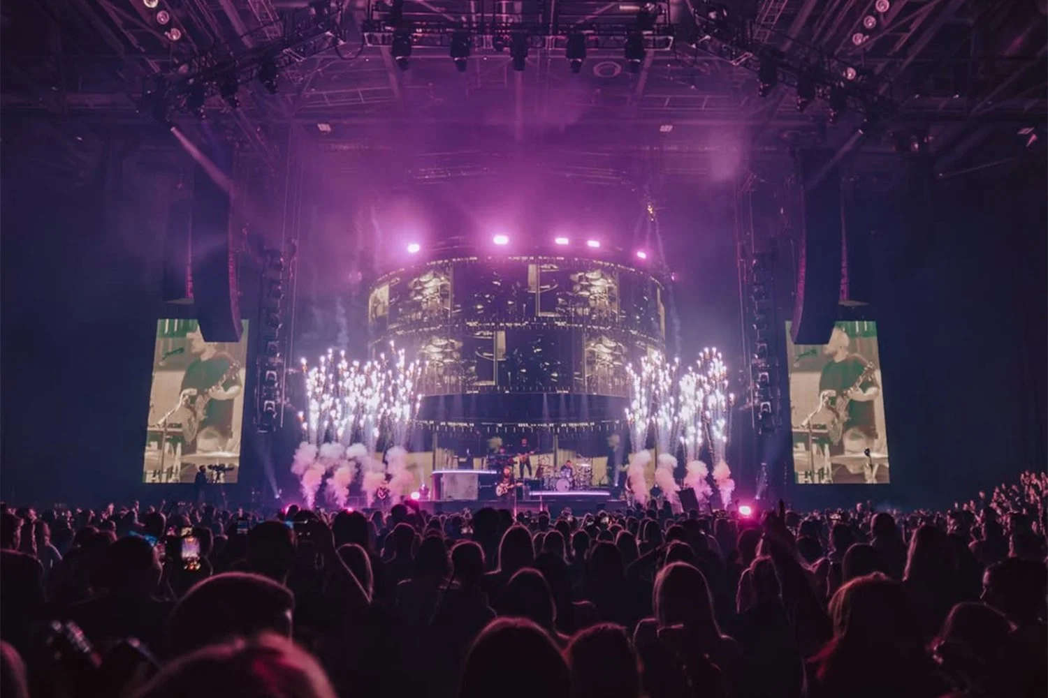 Concert stage with bright pink and purple lighting, large screens showing a guitarist, fireworks and smoke effects, and a crowded audience in a dark indoor arena.