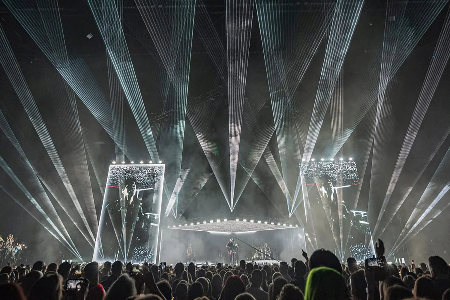 Concert stage with bright lights, large screen displays, and audience raising hands inside a dark indoor venue.