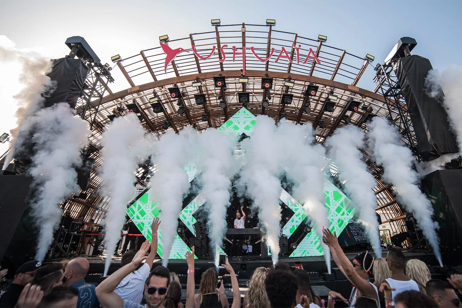 An outdoor music festival stage with a DJ performing, surrounded by smoke effects, digital screens displaying green graphics, and a crowd of attendees in front.