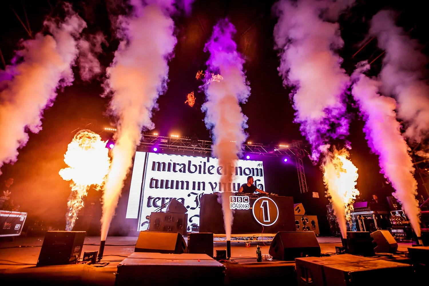 A DJ performs on stage at BBC Radio 1 event, with large flames and colorful smoke effects, and a large screen displaying visuals and the BBC Radio 1 logo.