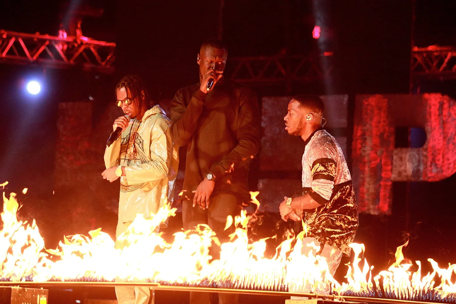 Three male performers on stage surrounded by flames with a dark background and stage lighting.
