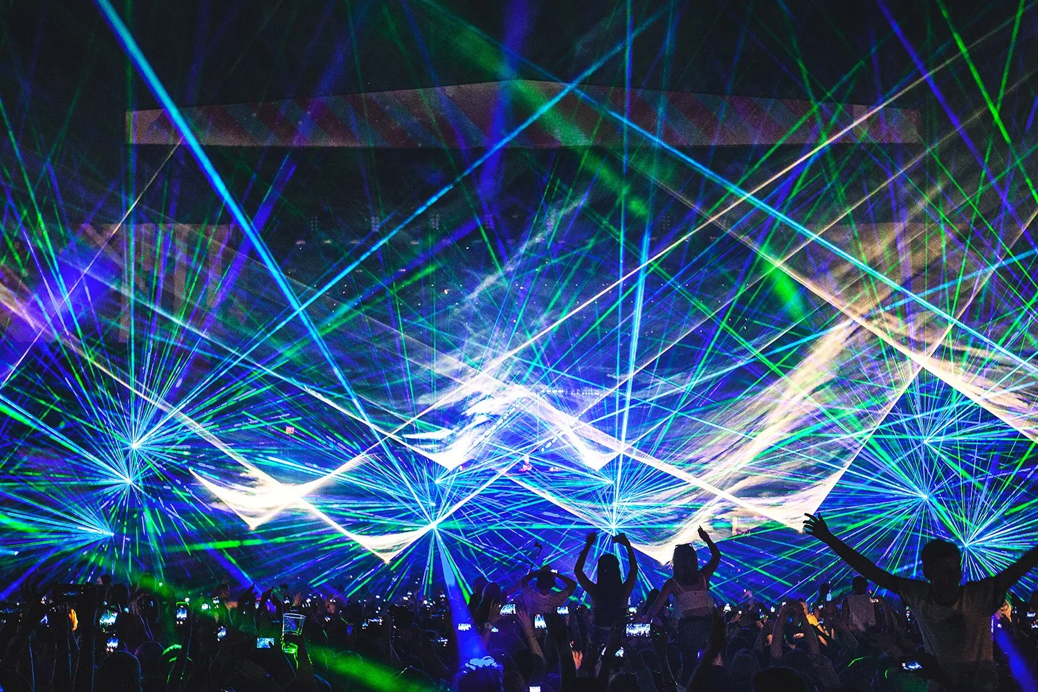 A concert or dance event with colorful laser lights and a crowd of people, some dancing and taking photos.