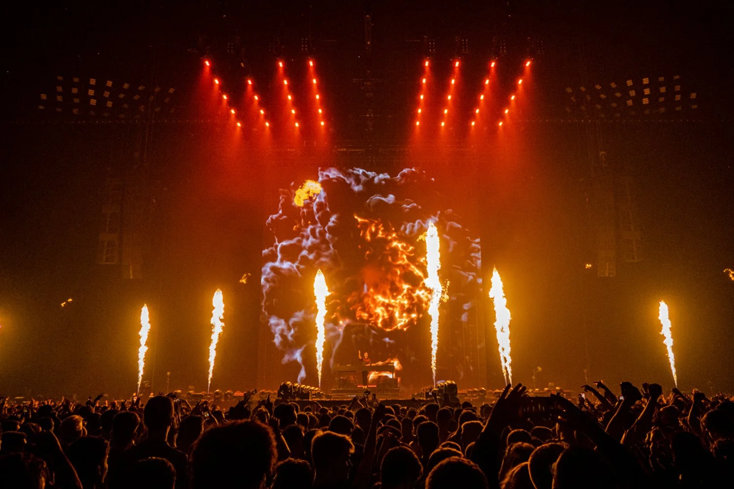 Concert stage with fire and smoke effects, fireworks, a performer at a keyboard, and an audience in the foreground.
