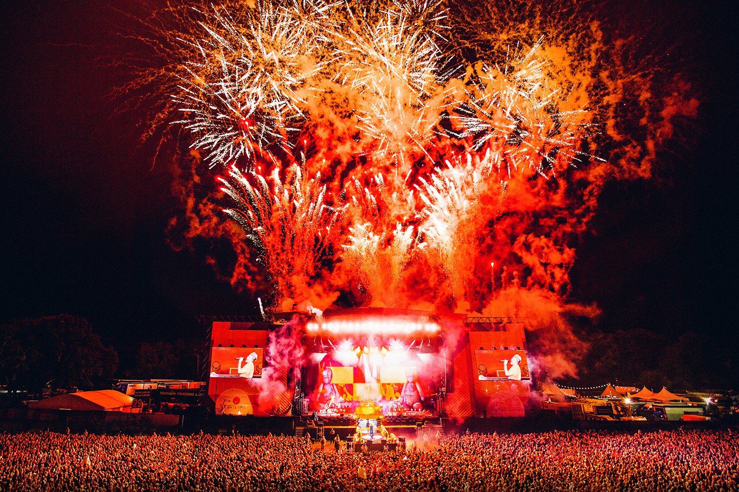 A large crowd gathered at an outdoor concert during fireworks display at night, with colorful fireworks lighting up the sky and stage.