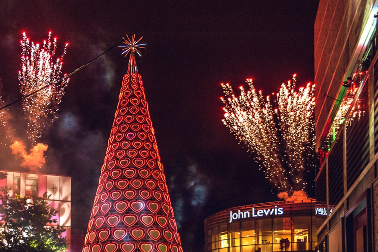 Nighttime city scene with a Christmas tree decorated with red heart-shaped lights, surrounded by fireworks exploding in the sky, and a building with a sign that reads 'John Lewis'.