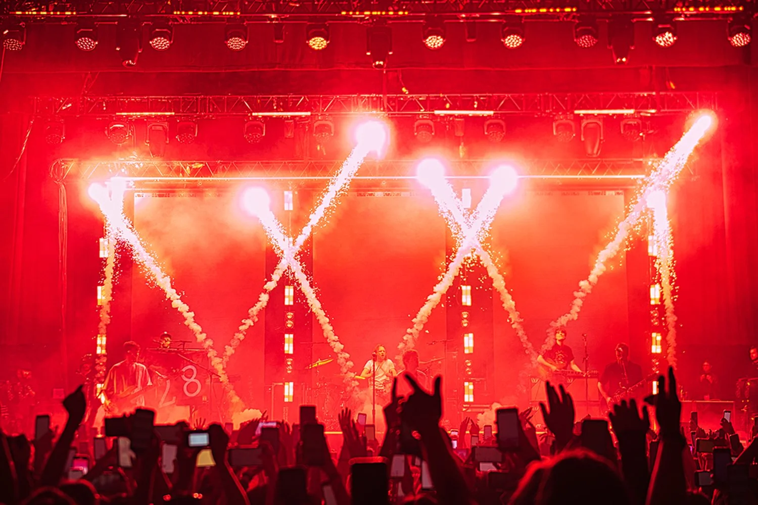 Concert stage illuminated with bright red lighting and pyrotechnics, with band members performing and audience in silhouette raising hands.