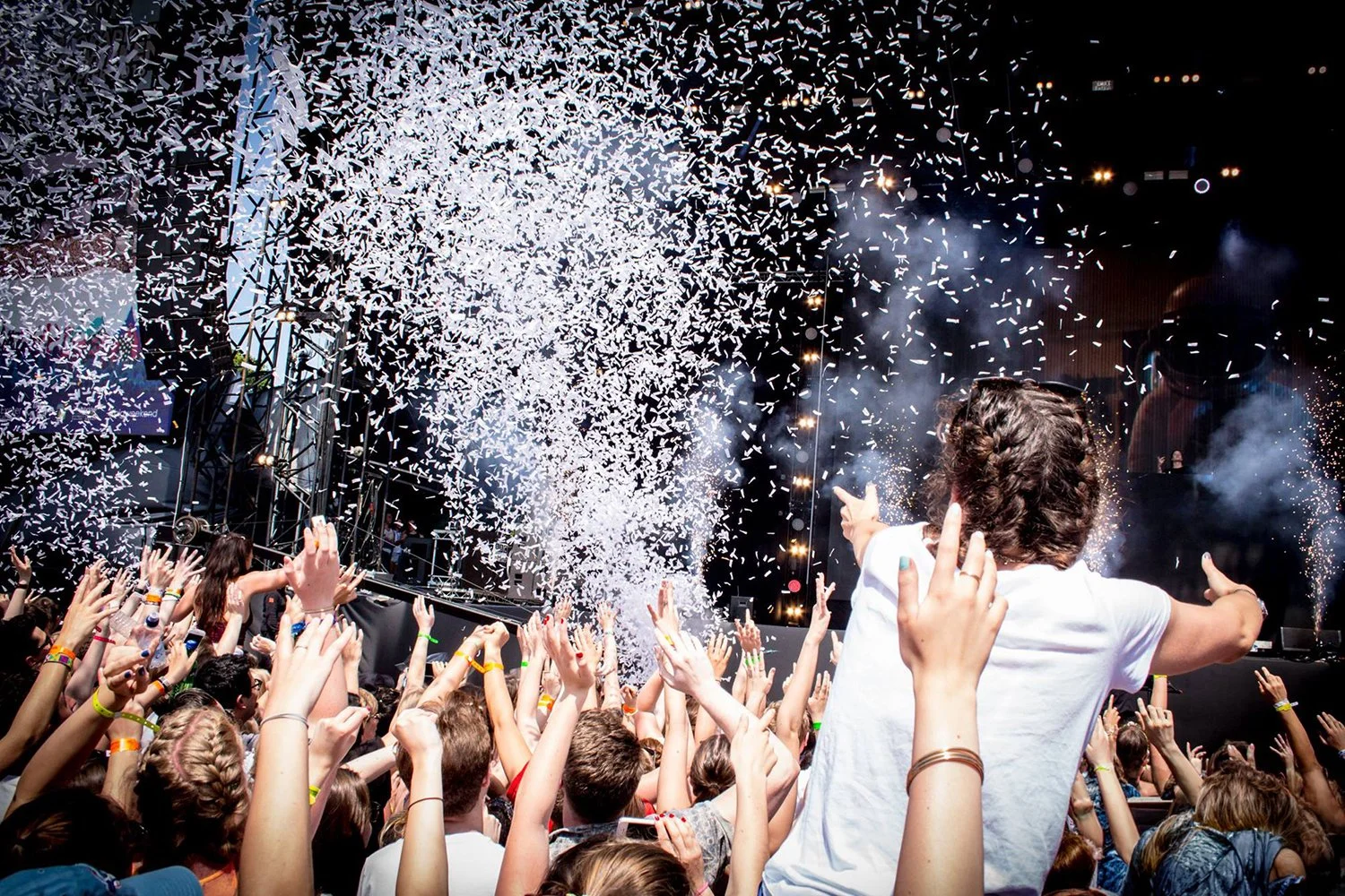 Crowd at a concert or festival celebrating with confetti and fireworks, people raising their hands, stage with lights in the background.