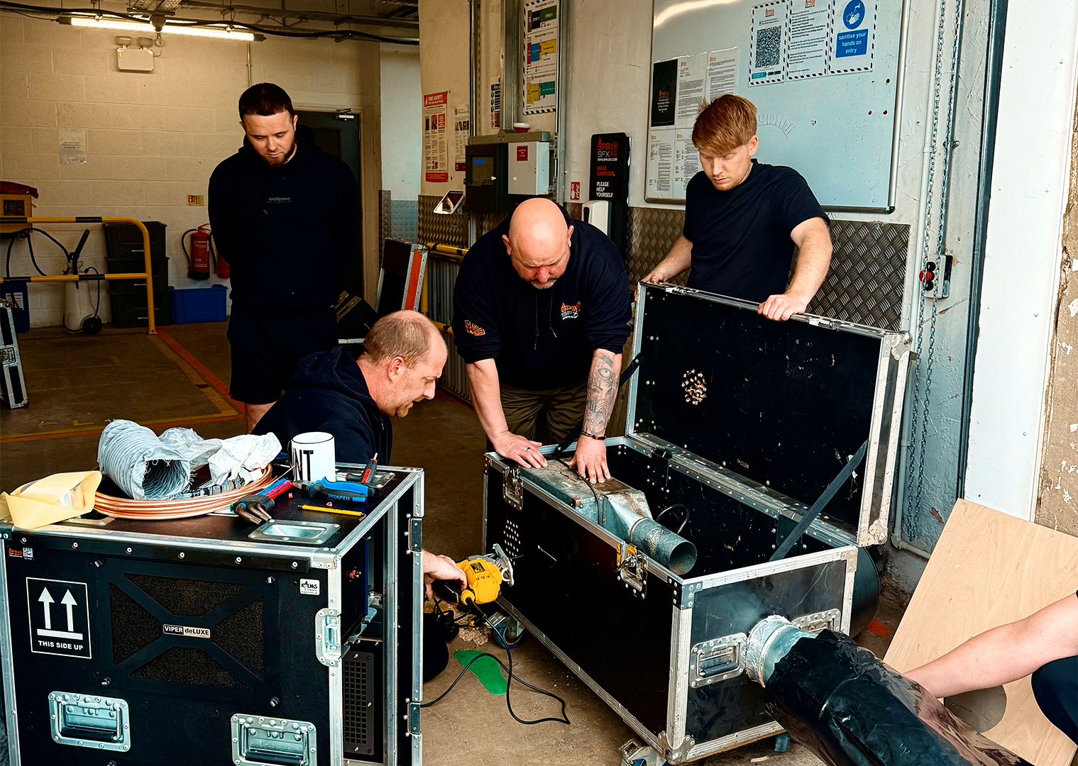 Five men working together on a technical project in an industrial setting, with various tools, equipment, and a large black mechanical device or machine.