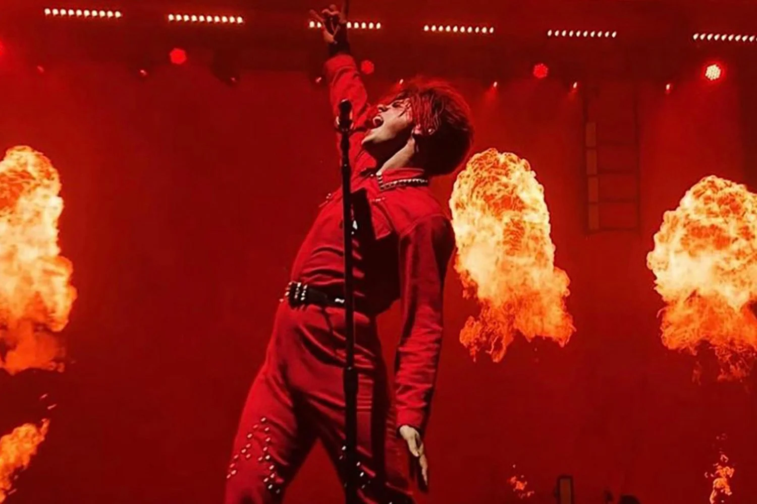 performer singing on stage with flames and fire effects in the background, dressed in a red outfit, head tilted back.