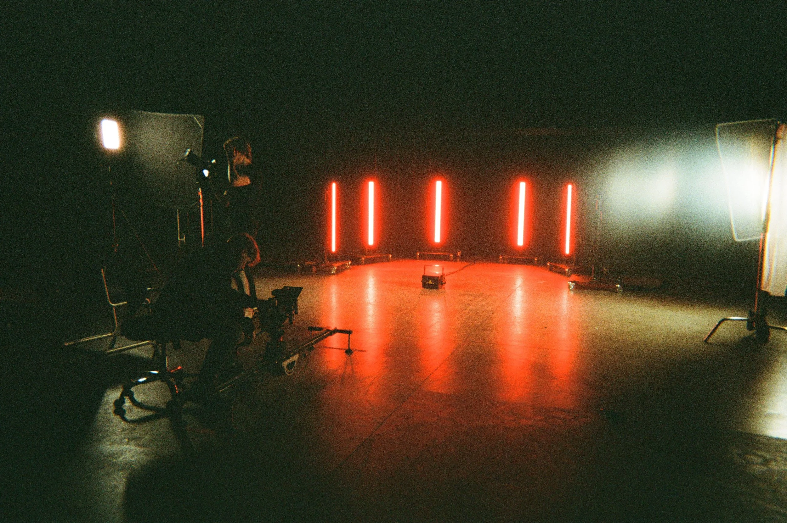Dimly lit indoor stage with five red vertical lights, two large white fabric screens on the sides, and a person setting up equipment.