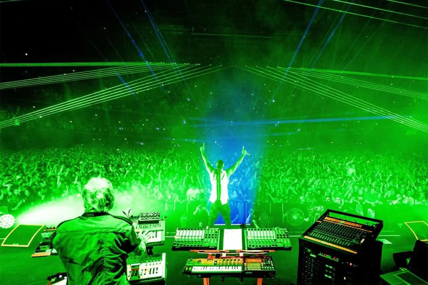 A DJ performs at a concert with a large crowd, green and blue laser lights shoot across the stage, and the DJ and audience are illuminated by vibrant lighting.