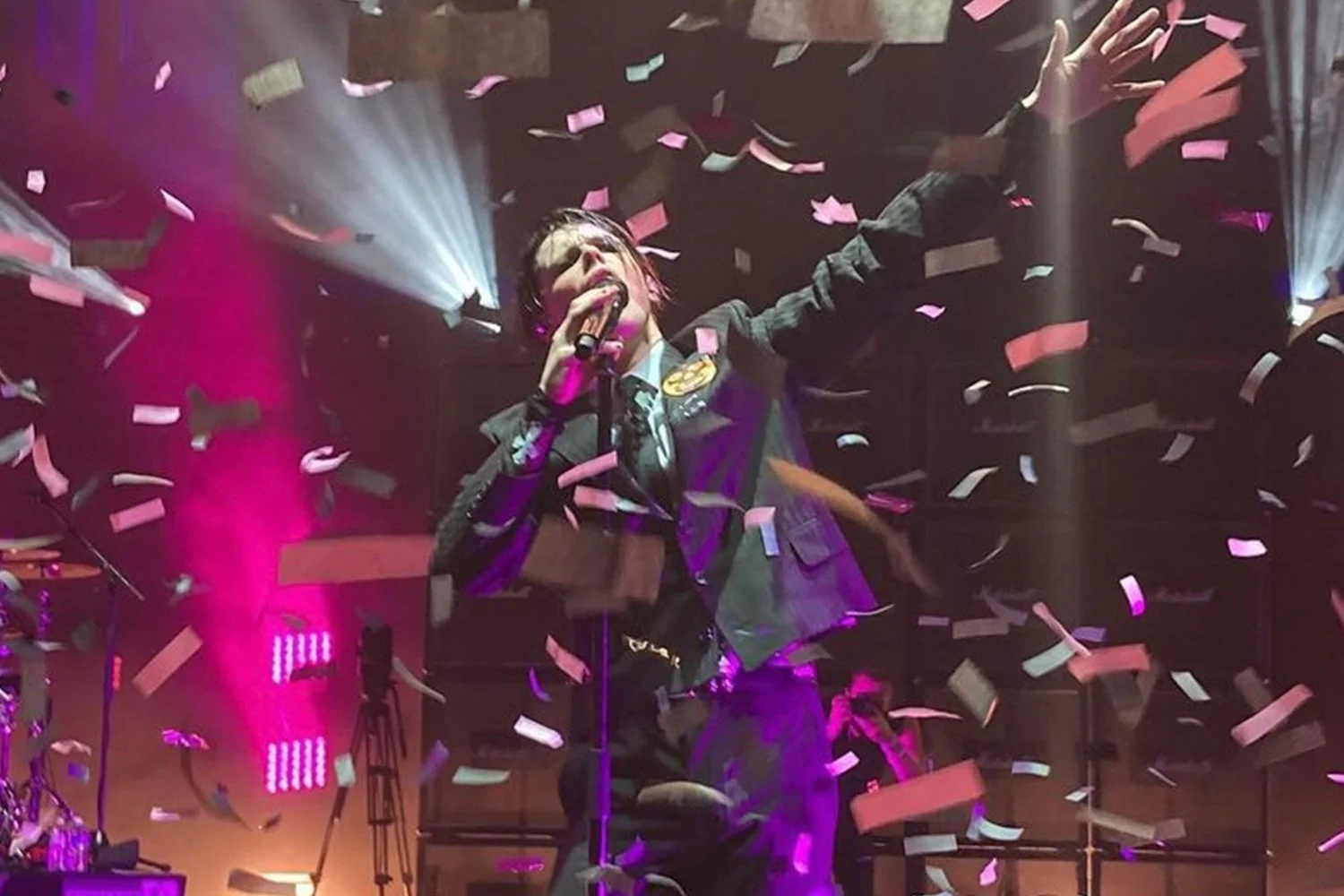 Performer singing into a microphone on stage, surrounded by falling confetti and colorful stage lights.