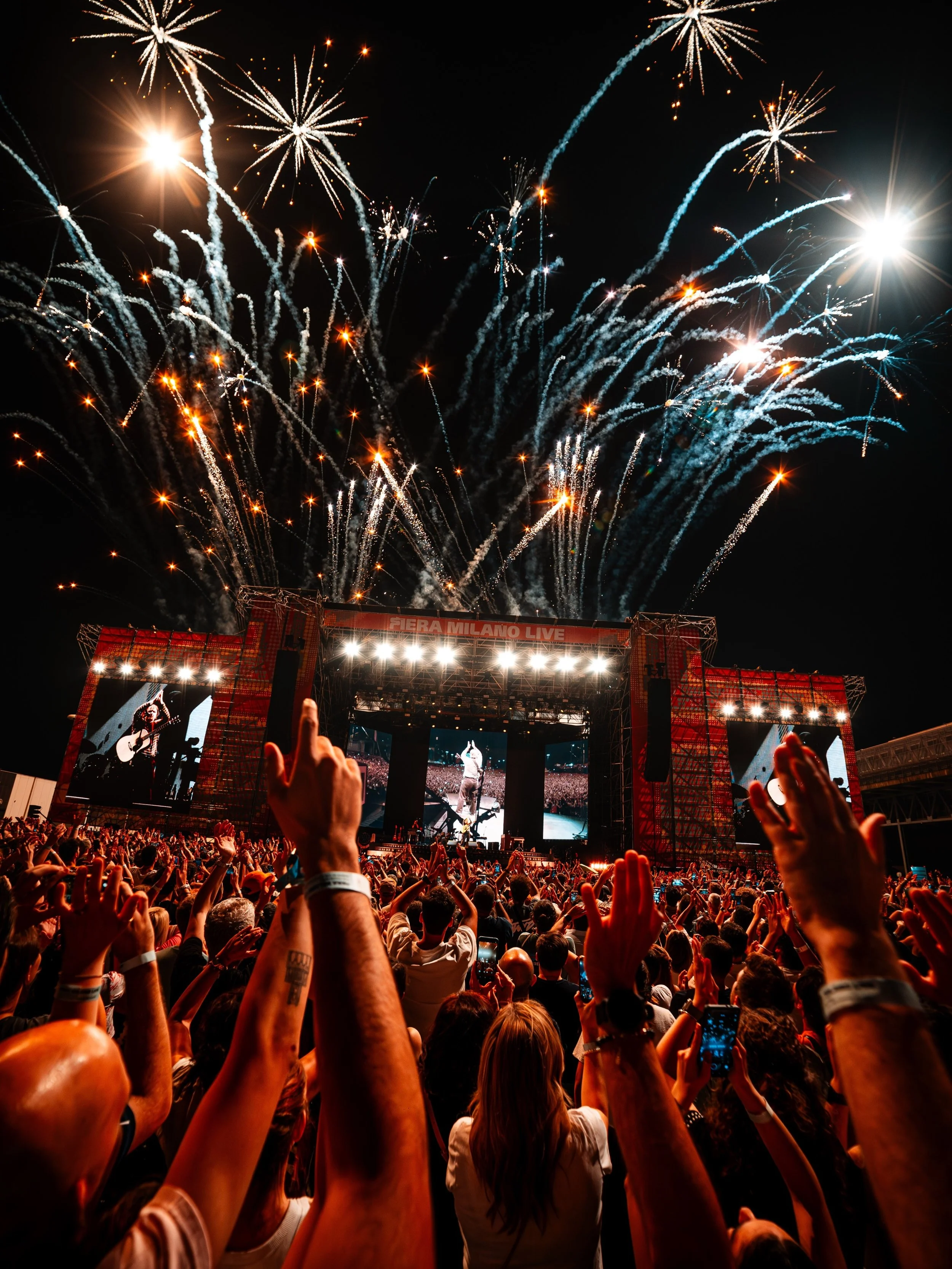 Crowd at a concert with fireworks exploding in the sky at night, stage with screens, and audience raising hands.