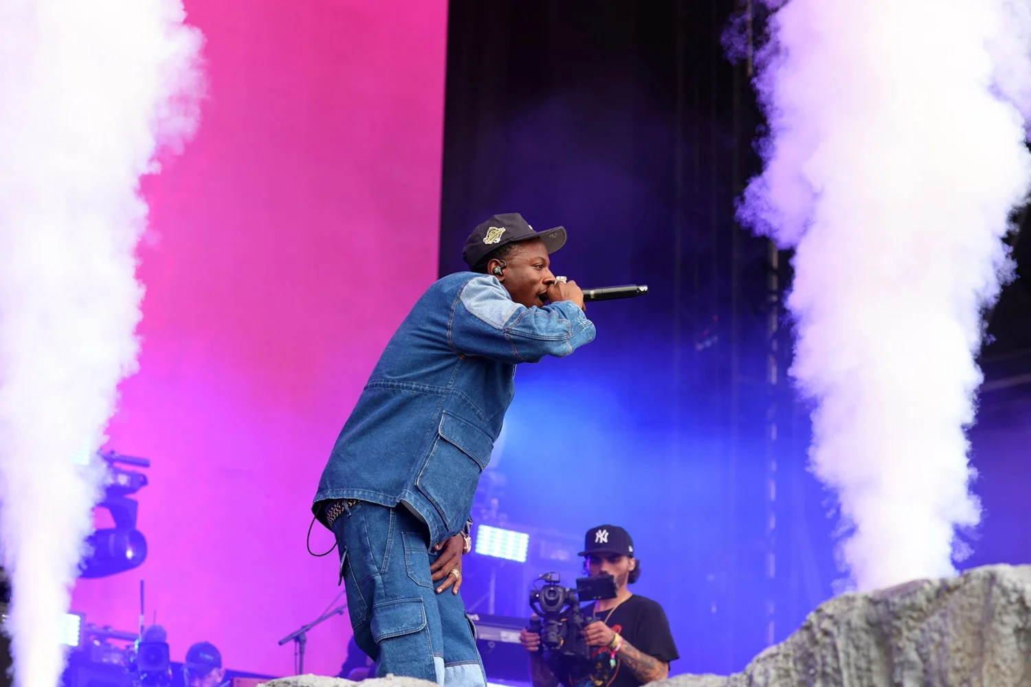 A performer in a denim jacket and cap singing into a microphone on stage with colorful lights and smoke effects.