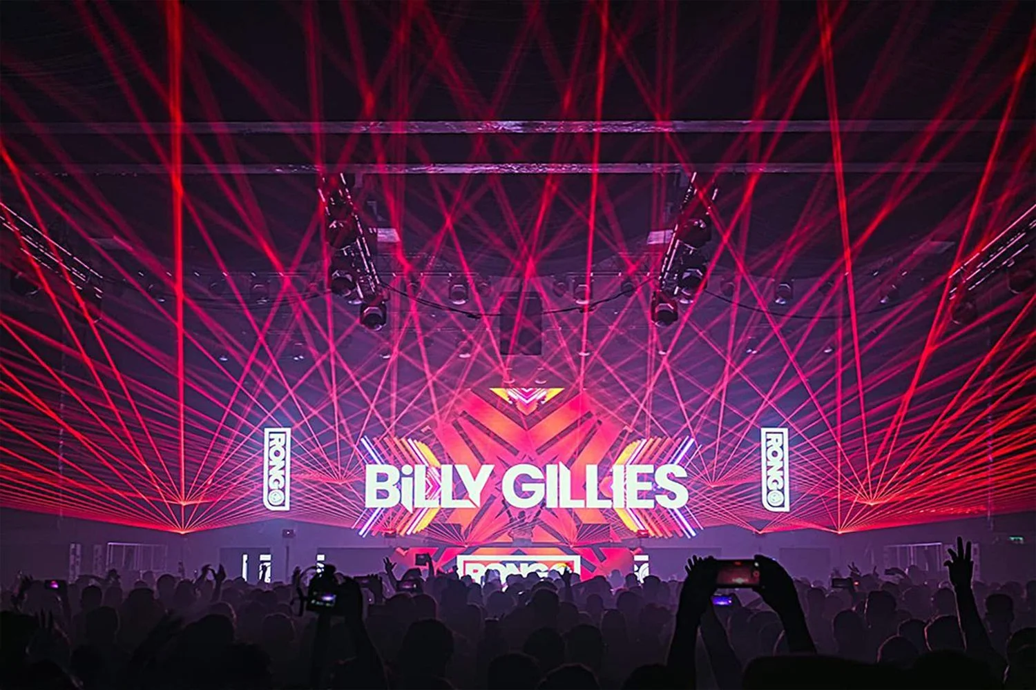Concert stage with red laser lights, large screen displaying "BILLY GILLIES," and silhouetted audience holding up phones.