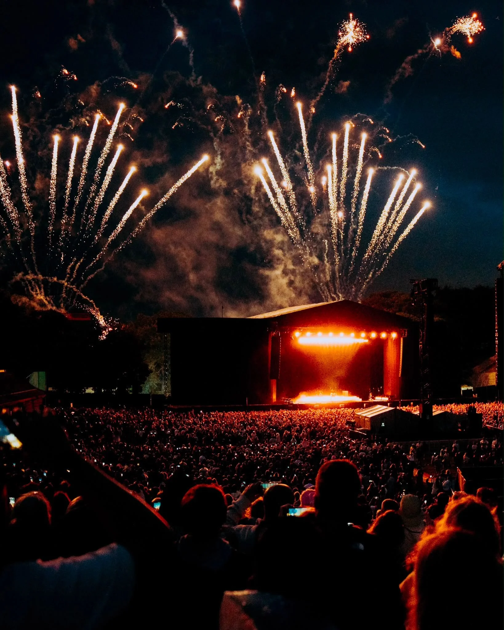 Nighttime scene of a large crowd at an outdoor concert or festival, with fireworks lighting up the sky above the stage, which is illuminated with orange lights.