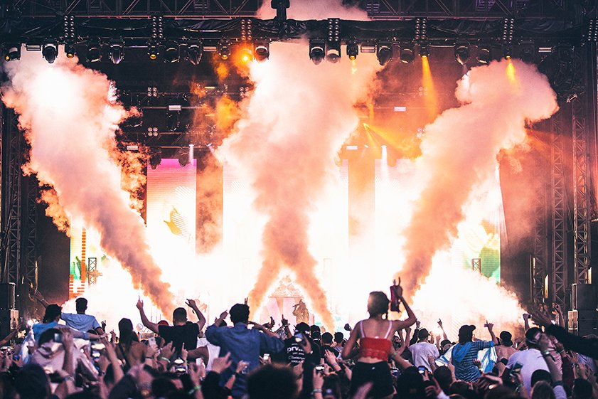 Crowd at concert or festival with stage lights and smoke effects in the background.