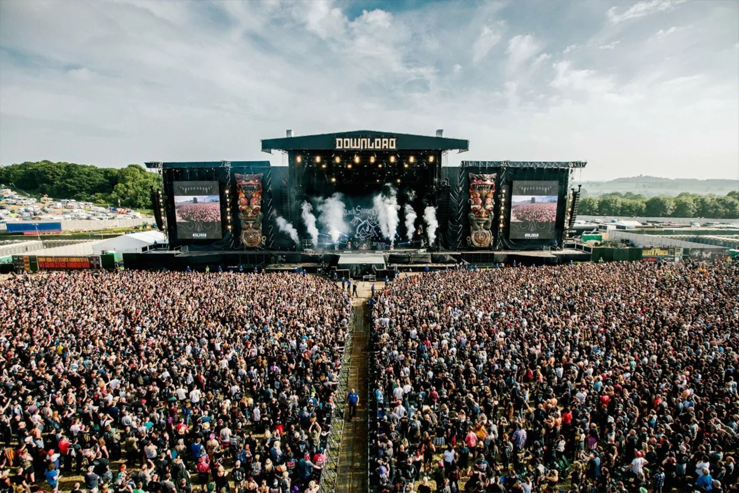A large outdoor music festival with a massive crowd facing a big stage with smoke effects, two large screens, and the word 'DOWNLOAD' at the top.