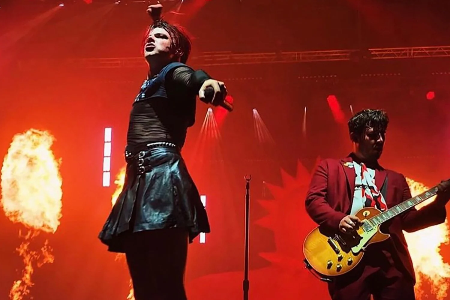 Two performers on stage with fire and red lighting behind them, one holding a microphone and pointing toward the camera, the other playing an electric guitar.