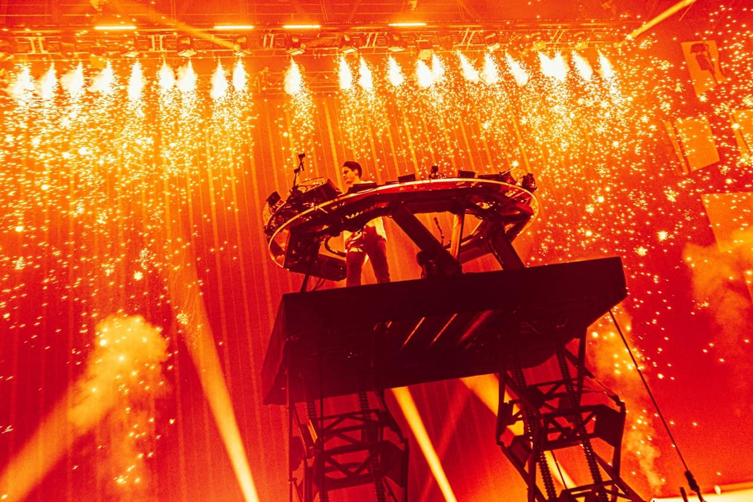A DJ performs on a platform surrounded by bright sparks and fireworks during a concert or festival, with vibrant orange and yellow lighting effects.