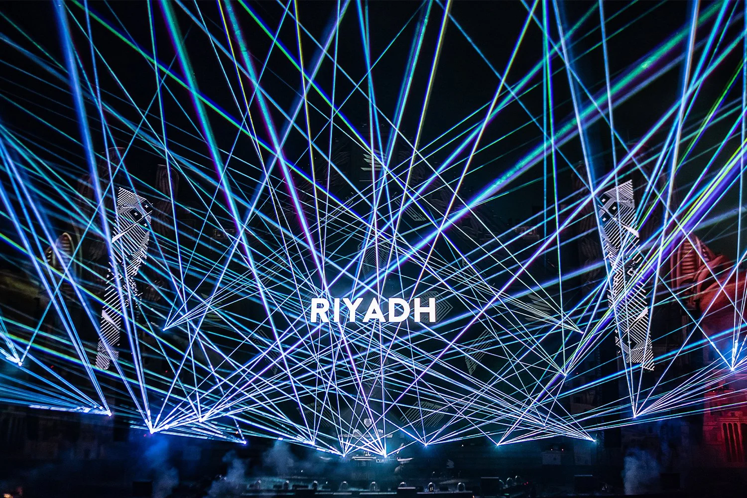 Nighttime laser light show with blue, green, and purple beams crisscrossing, displaying the word 'RIYADH' in the center.