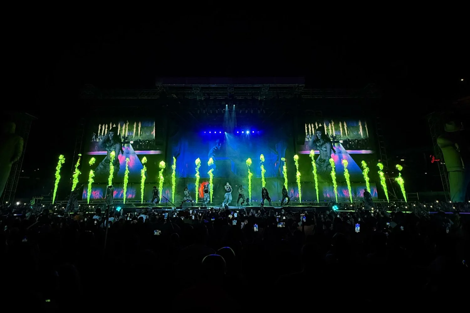 A stage performance with dancers, large screens displaying visuals, and green pyrotechnics in front of an audience holding up phones.