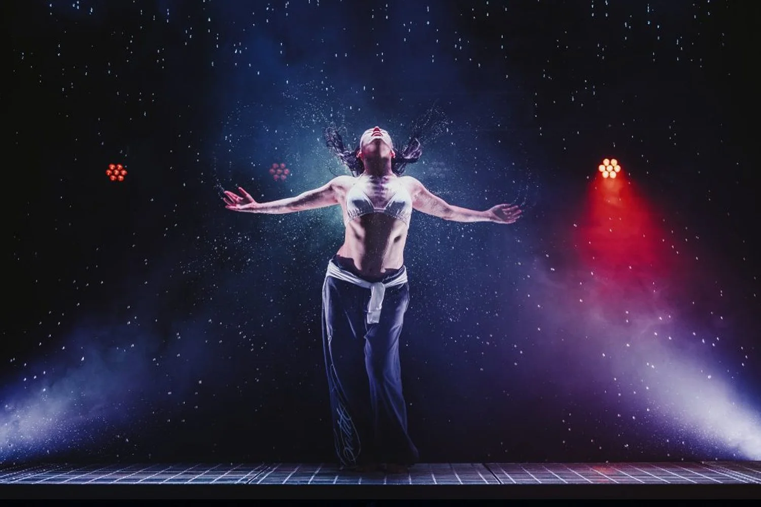 A performer in a bra and loose pants, standing on a stage with arms outstretched, with water splashing around her head and a starry background with stage lights.