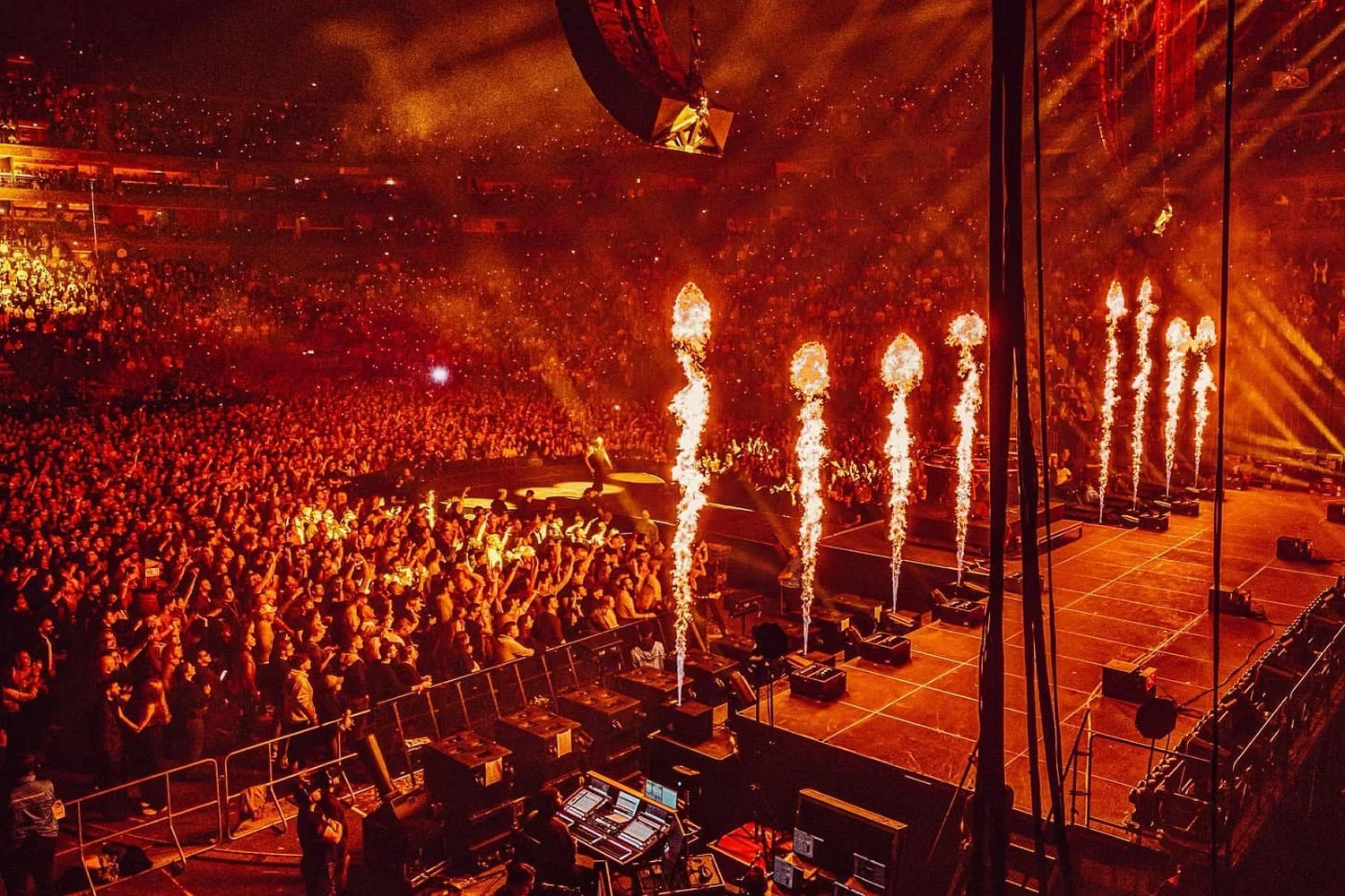 A concert scene with a large crowd and pyrotechnics firing on stage, with bright orange and yellow flames and sparks illuminating the audience.