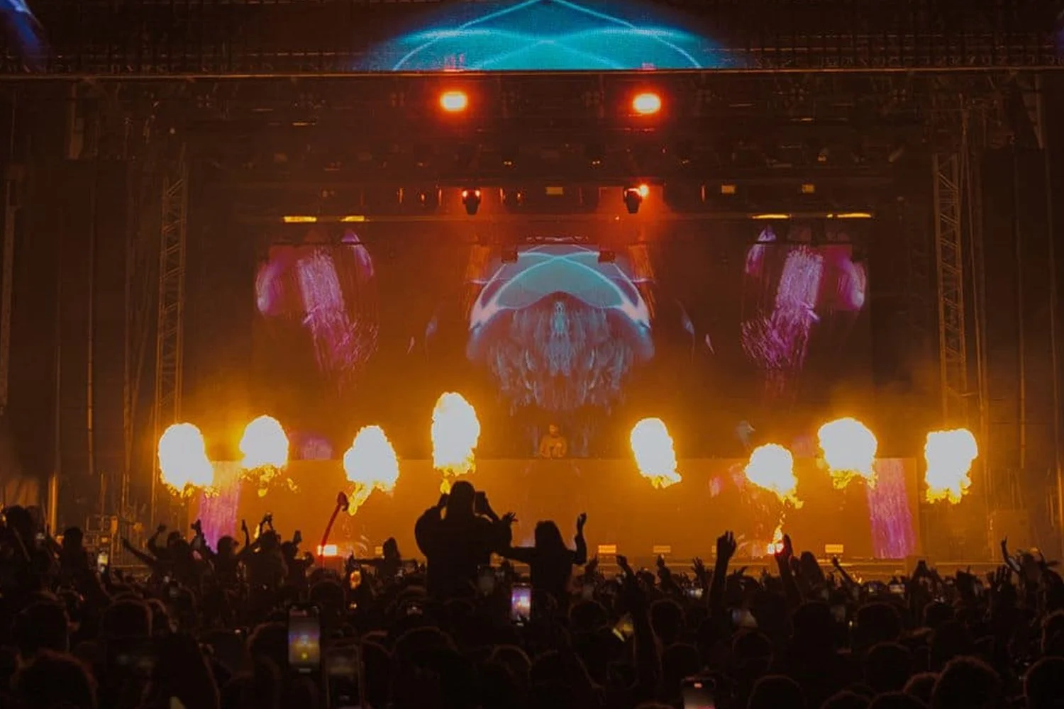 A live concert with a large audience, stage lighting, fire effects, and a digital screen displaying abstract visuals.