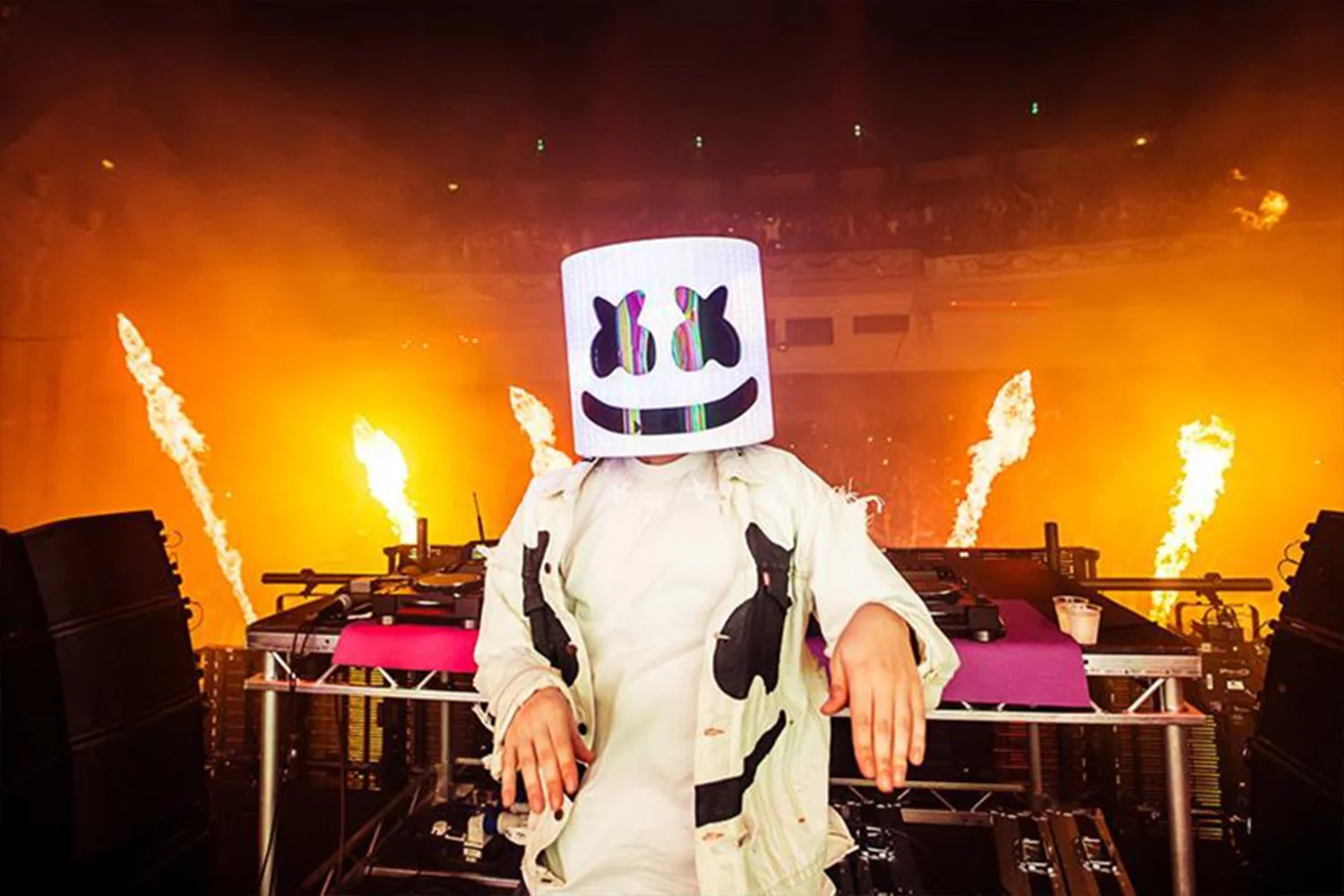 A DJ wearing a marshmello helmet and white clothing, standing behind a DJ booth with flames shooting in the background at a concert.