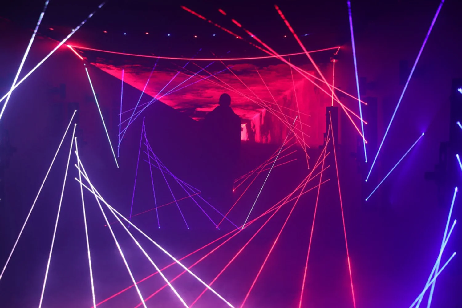 A person standing in front of a large LED screen displaying a red sunset or landscape scene, surrounded by dynamic laser light beams in red, blue, and white, creating an energetic visual display.