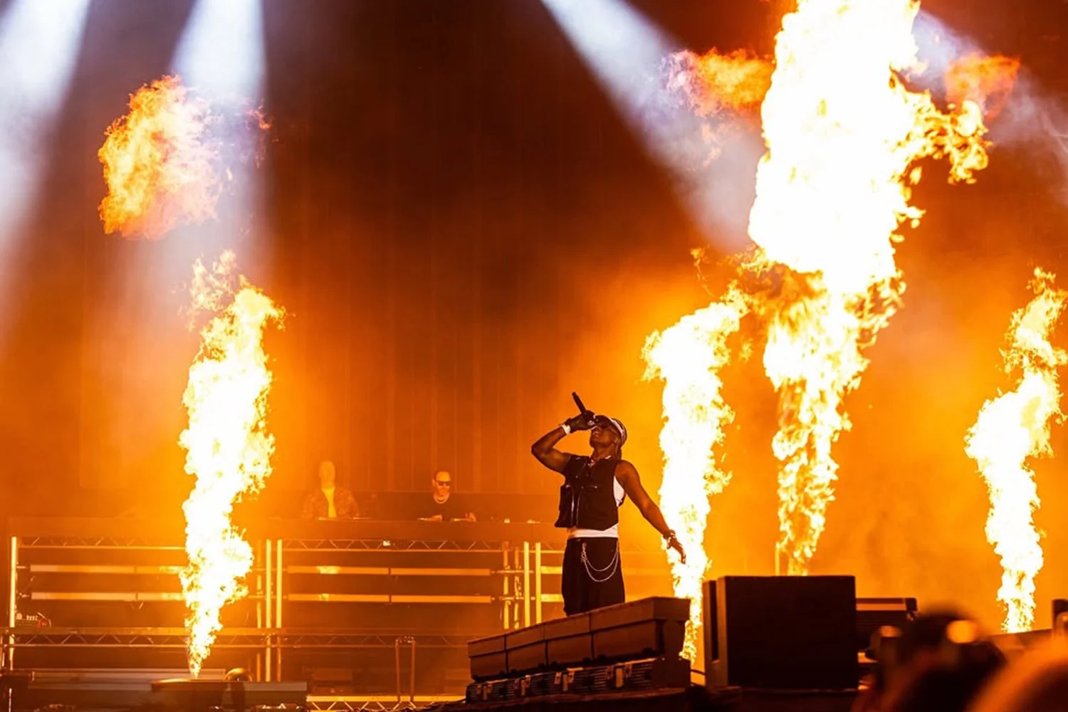 Performer on stage at a concert surrounded by flames and fire effects, with two DJs in the background.
