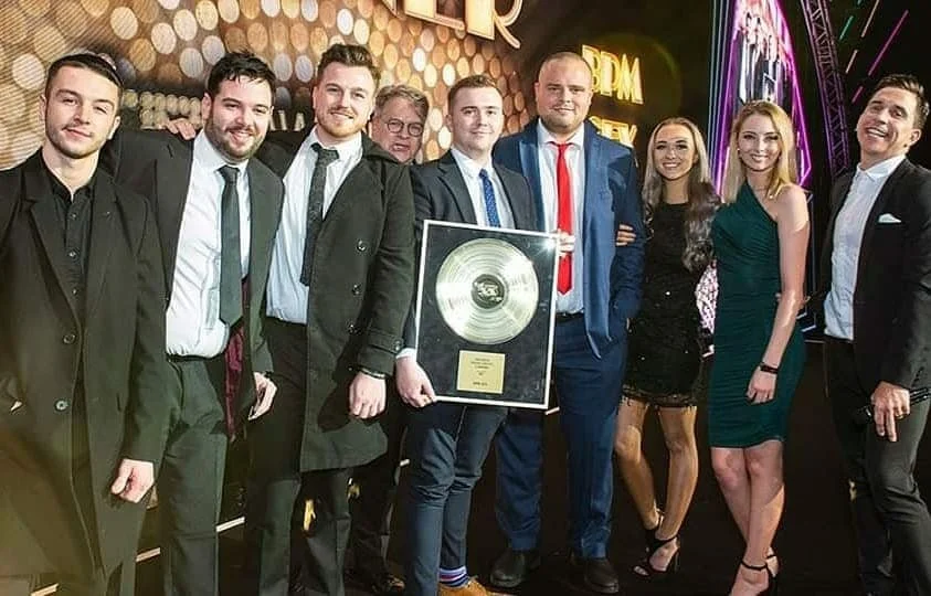 Group of nine people dressed in formal attire, celebrating an award in an event hall with gold and neon decor, with one person holding a large framed gold record.