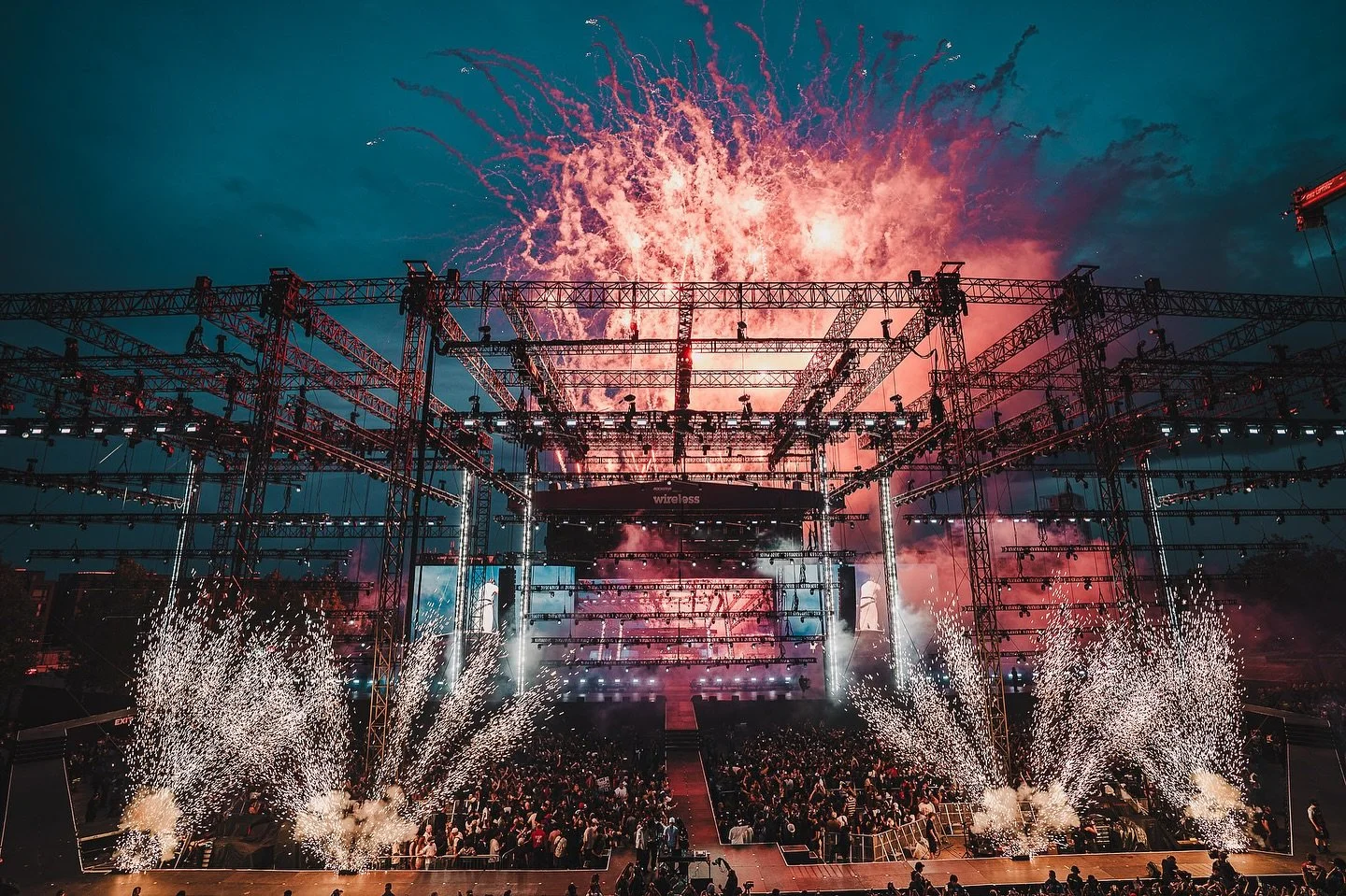 A large crowd at a concert or festival with fireworks and pyrotechnics over a stage with scaffolding and lighting structures.