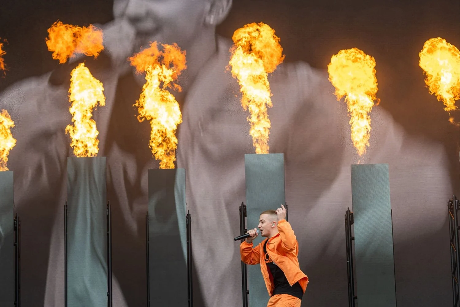 A performer in an orange outfit holding a microphone on stage with flames erupting from vertical panels behind them.