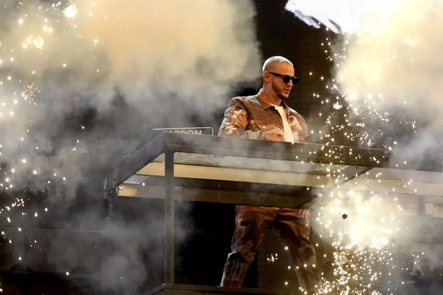 A man wearing sunglasses and camouflage clothing performing as a DJ behind a table surrounded by sparks and smoke.