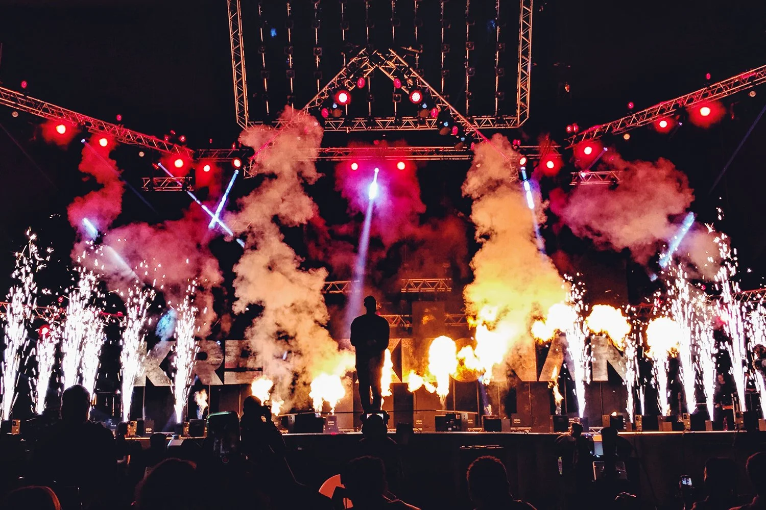 A concert stage at night with pyrotechnics, smoke, and colorful lights filled the scene. The silhouettes of audience members are visible in the foreground.