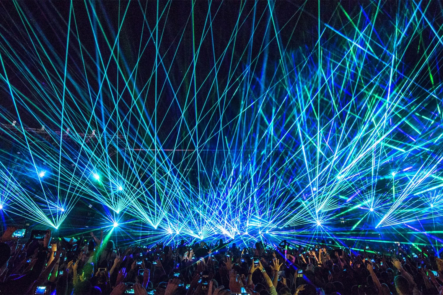 Bright blue and green laser lights at a concert or music event, with a large crowd raising their hands and taking photos.