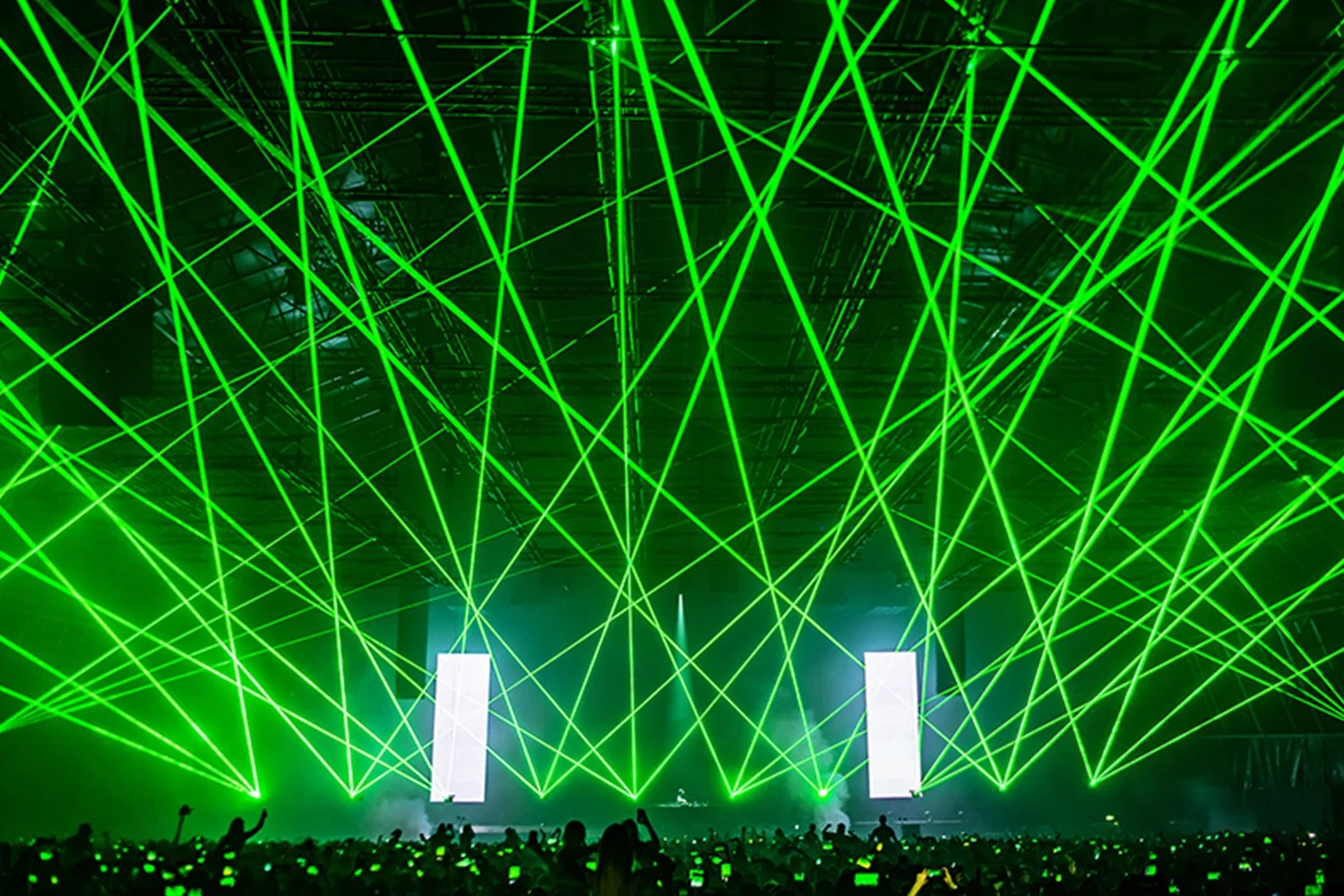 Concert or event with green laser lights crossing above a crowd of people with glow sticks, stage with two large screen displays.